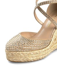 Beige espadrille with a rounded toe, embellished with clear rhinestones. Features a woven jute sole and crossed straps at the ankle.