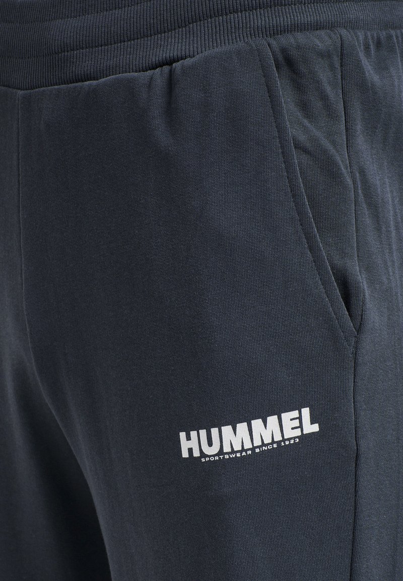 Pantalones de chándal marinos hechos de tela suave, con una cintura elástica, bolsillos laterales y un logo blanco de "HUMMEL" en el muslo izquierdo.