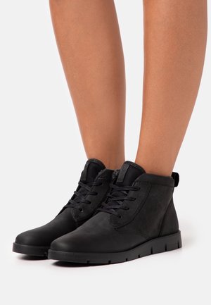 Bottines noires à lacets avec col rembourré portées sur des jambes nues sur fond blanc.