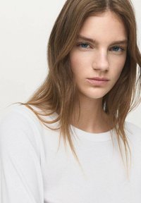 Cămașă albă cu mâneci lungi și guler rotund, purtată de o persoană cu păr lung, castaniu și machiaj natural, pe un fundal deschis.