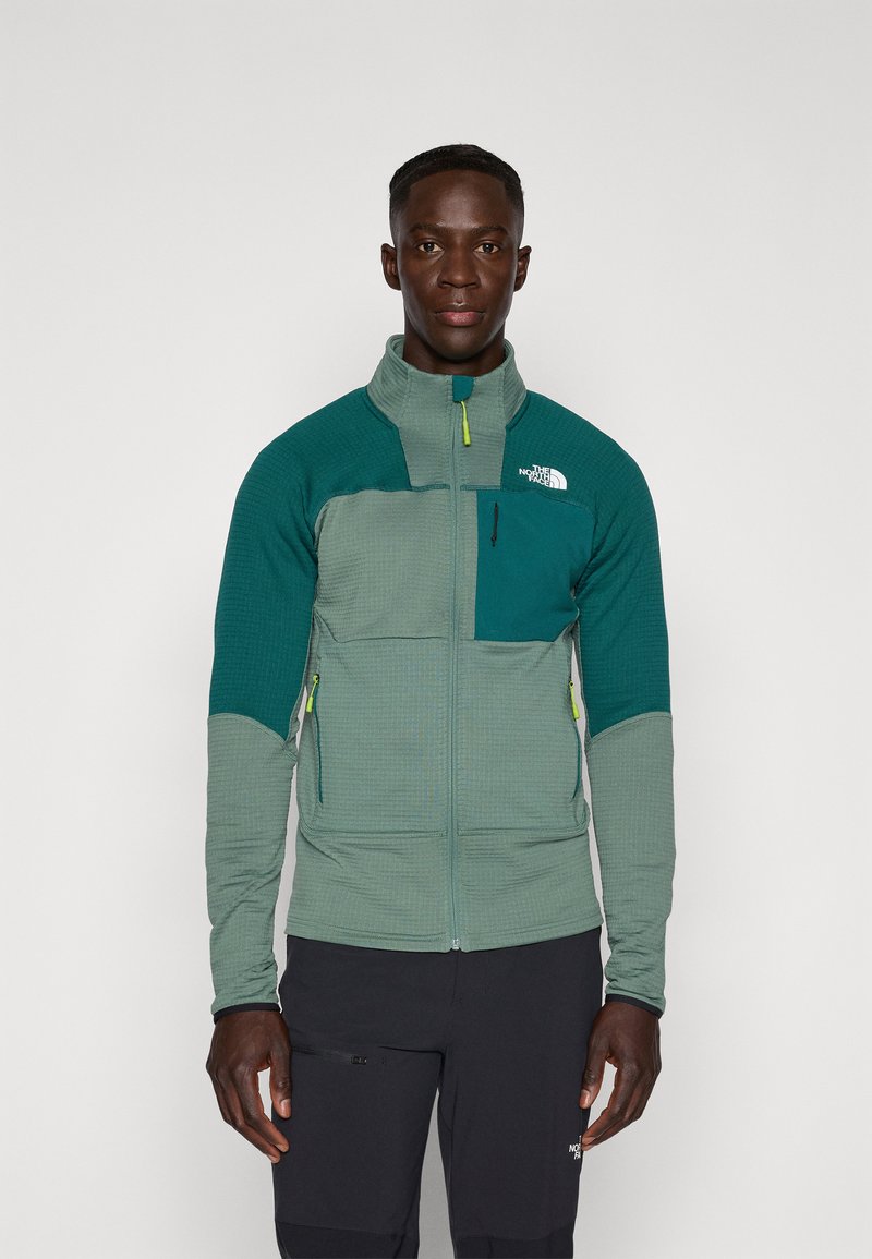 Green and teal fleece jacket with a high collar, textured pattern, zippered chest pocket, and contrasting zippers. Suitable for outdoor use.