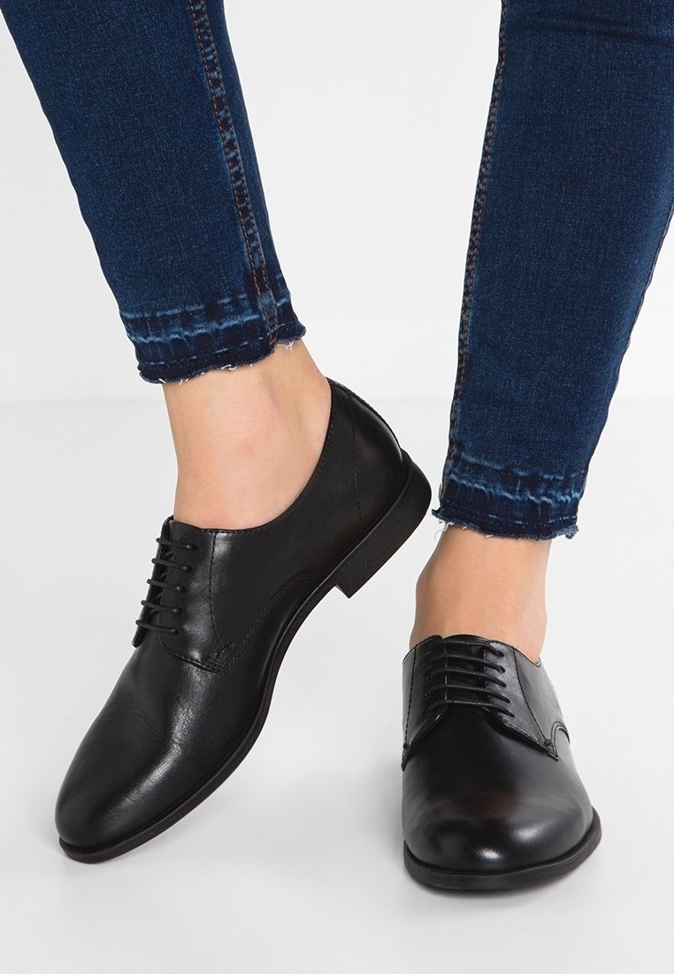 Chaussures Oxford en cuir noir portées avec un jean skinny bleu foncé à ourlet effiloché sur fond blanc.