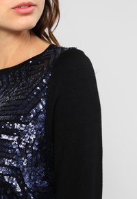 Haut du torse d'une femme portant un pull noir avec un motif de sequins géométriques bleu foncé, lèvres légèrement entrouvertes et cheveux bruns légèrement bouclés.