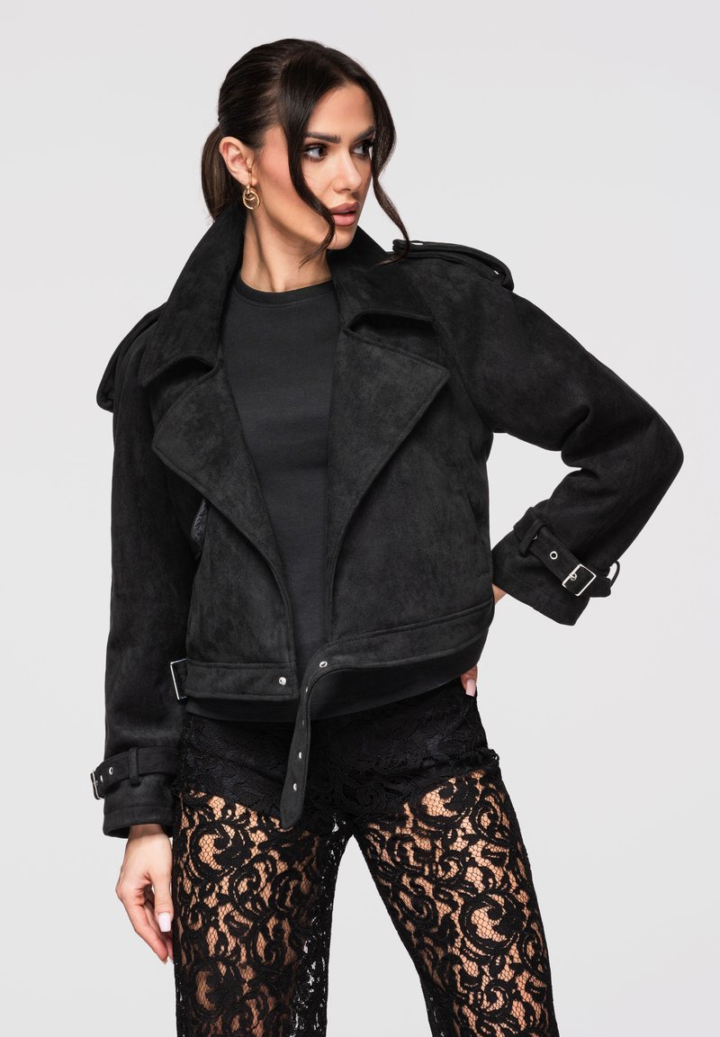 Femme portant une veste en daim noire avec des boucles et un pantalon transparent à motif de dentelle noire, regardant légèrement vers sa gauche.