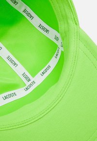 Lys grøn tekstureret stof med en rund halsudskæring og et hvidt stofmærke med "LACOSTE" trykt gentagne gange indeni.