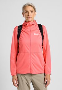 Koraal kleurige zip-up jack met een hoge kraag, rekbare stof, lange mouwen en logo-detail, gecombineerd met een zwarte rugzak en lichtgekleurde broek.