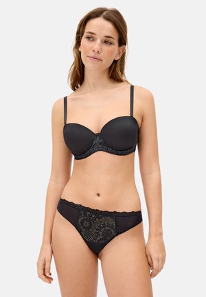 Mujer con conjunto de sujetador y bragas de encaje negro a juego, de pie con expresión neutral contra un fondo liso.