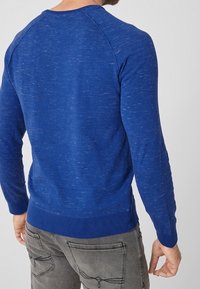 Maglione azzurro lavorato a maglia con finitura testurizzata, maniche raglan e orlo a coste. La vista posteriore mostra un design aderente. Abbinato a jeans grigi chiari.