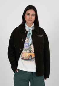 Frau trägt eine dunkle Cordjacke, ein T-Shirt mit einem Vintage-Auto, türkisfarbene Hosen und einen hellblauen gemusterten Schal um den Hals.