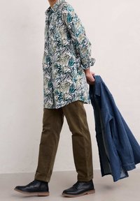 Langärmliges, kragenloses Hemd mit grün-blauem botanischem Druck, kombiniert mit olivgrünen Hosen und schwarzen Stiefeln, hält eine Jeansjacke.