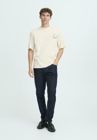 Beige kortærmet t-shirt med en grafik på venstre bryst, parret med mørkeblue jeans og sorte sko, båret af en stående model.