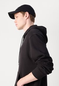Sudadera negra con capucha y bolsillo tipo canguro, tejido texturizado y puños acanalados, complementada con una gorra negra a juego, vista de lado.