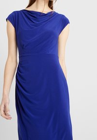 Abito blu royal con scollatura drappeggiata, maniche a cappuccio e dettaglio laterale arricciato, realizzato in tessuto liscio ed elasticizzato.