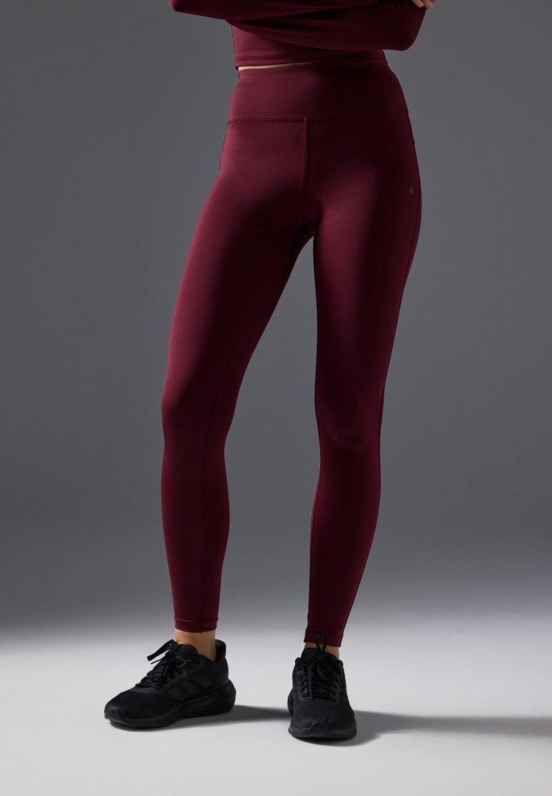 Burgundy leggings made of stretch fabric, high-waisted design, smooth texture, paired with black athletic shoes.