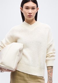 Νεαρός με ίσια σκουρόχρωμα μαλλιά φοράει εκρού πουλόβερ πλεχτό και ελιές παντελόνι, κρατώντας μια μεγάλη μαλακή λευκή τσάντα, δείχνοντας το τατουάζ στο αντιβράχιο.