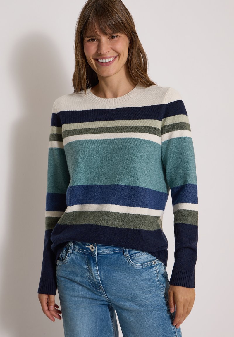 Femme souriante portant un pull rayé bleu, vert, blanc et marine à manches longues et un jean bleu clair sur un fond uni.