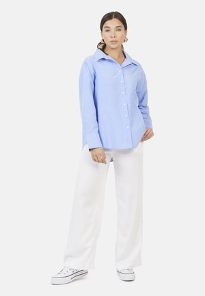 Donna in piedi che indossa una camicia azzurra a bottoni, pantaloni bianchi a gamba larga, sneakers bianche e orecchini a cerchio dorati su sfondo bianco.