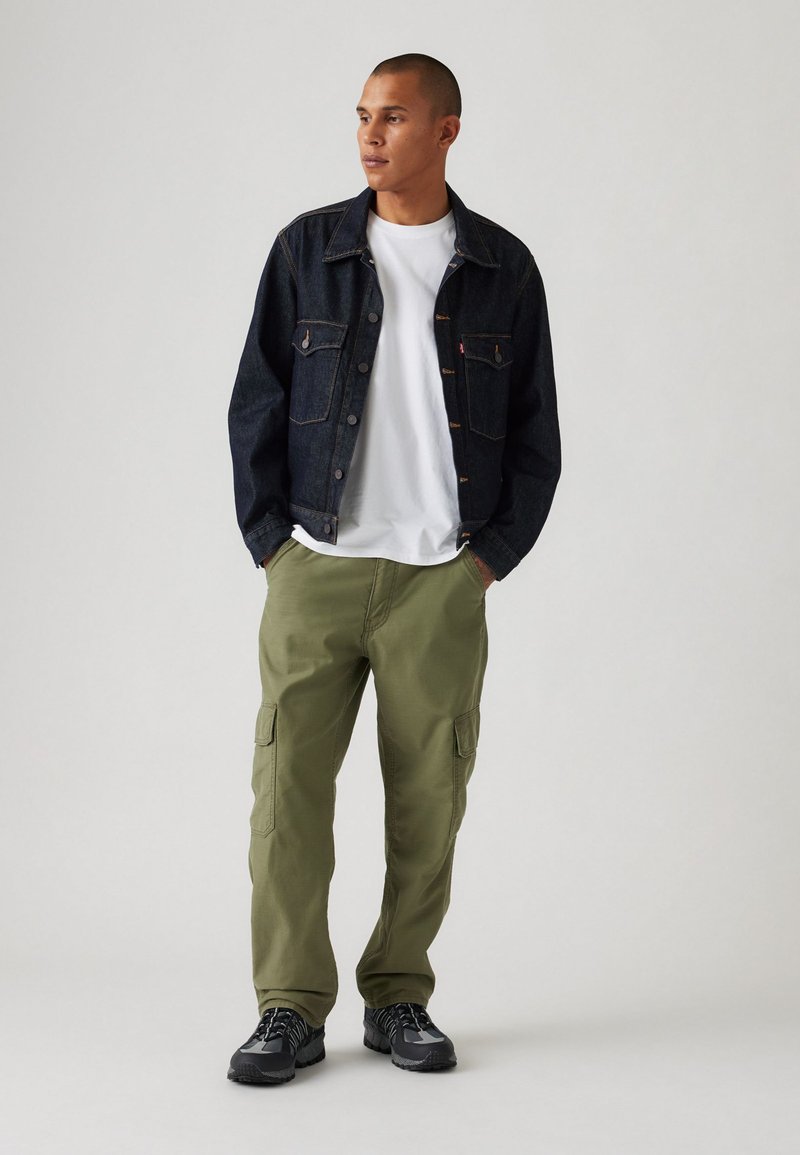 Giacca di jeans con tasche frontali, t-shirt bianca, pantaloni cargo verde oliva e sneakers nere. Vestibilità casual e stile utilitario.