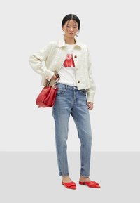 Chaqueta blanca con lentejuelas, camiseta gráfica roja, vaqueros de mezclilla azul claro y zapatos planos rojos. La modelo sostiene un bolso rojo con detalle de cadena.