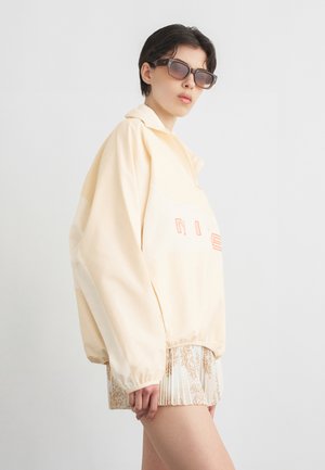 Jeune personne portant une veste beige oversize avec un texte corail, des lunettes de soleil marron et un short plissé à motifs, debout devant un fond uni.