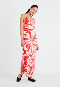 Vestido maxi en estampado floral rojo y blanco, con tirantes de espagueti, aberturas laterales y una tela ligera. Acentuado con zapatos negros de tacón redondo.