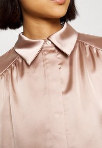 Blouse en satin de couleur rose pâle avec un col pointu, un éclat subtil et des détails froncés aux épaules. Fermeture par un bouton unique au niveau du cou.