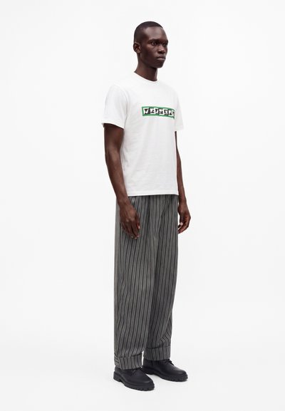 Homme portant une chemise blanche à manches courtes avec un texte graphique et un pantalon gris ample rayé avec des chaussures noires, debout, faisant face légèrement vers la droite.