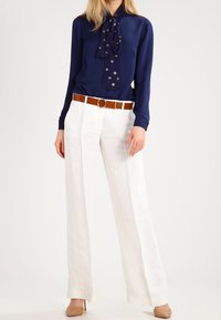 Blouse marine avec un détail de col noué, pantalon blanc à jambes larges et une ceinture marron avec une boucle ronde. Tissu doux et coupe ajustée.