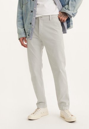 Uomo che indossa pantaloni grigio chiaro a gamba dritta, scarpe da ginnastica bianche, camicia bianca e giacca di jeans, in piedi davanti a uno sfondo semplice.