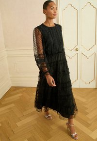 Rochie neagră cu buline și tull volanf, mâneci lungi transparente, talie evidențiată și tiv în straturi. Asortată cu sandale metalizate cu bretele.