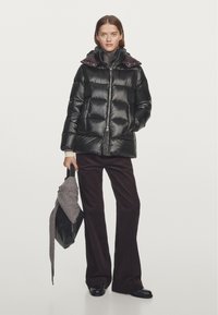 Giacca imbottita nera con finitura lucida, chiusura a zip e colletto viola a contrasto. Abbinata a pantaloni larghi scuri e una borsa nera.