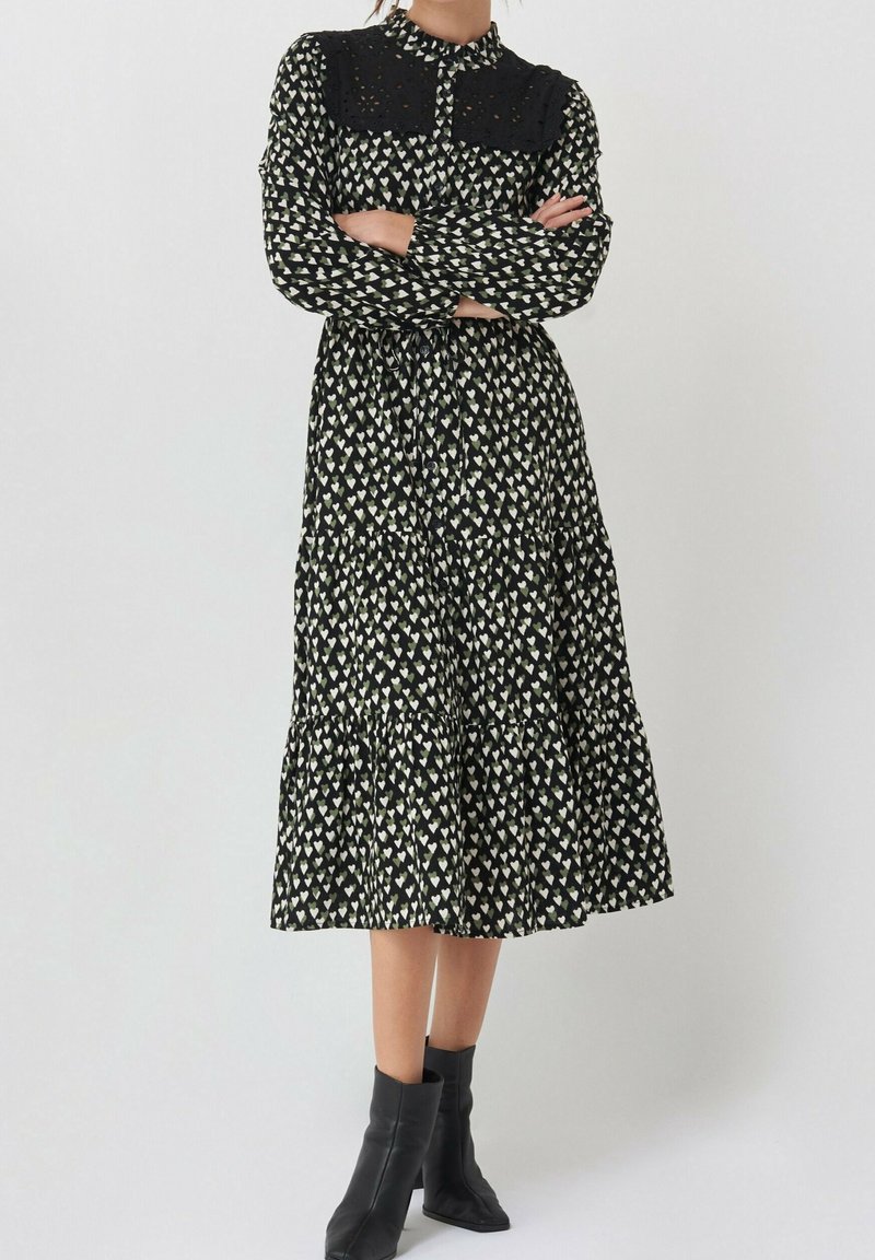 Femme portant une robe midi noire à motif de cœurs blancs, manches longues, empiècement en dentelle et bottines noires, debout les bras croisés.