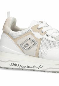 Baskets blanches avec des accents beiges, matériau en maille, détail du logo argenté, panneaux texturés et une semelle blanche épaisse avec une bande de roulement noire.