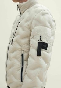 Puffige weiße Jacke mit hohem Kragen, diagonaler Steppung, Reißverschlusstaschen und einem schwarzen Etikett am Ärmel.