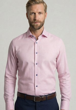 Blonde man met baard, gekleed in een lichtroze overhemd met knopen, ingestopt in een donkere broek met een bruine riem, staand tegen een effen achtergrond.