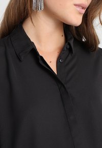 Camicia nera con colletto realizzata in tessuto liscio, dotata di chiusura a bottoni. Abbinata a orecchini in argento testurizzati per un tocco di stile.