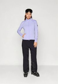 Kobieta w lilakowej kurtce z zamkiem, czarnych spodniach narciarskich, butach narciarskich i czarnych goglach narciarskich na głowie, na jednolitym tle.