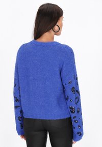 Zils adīts džemperis ar garām piedurknēm, kas rotāts ar melnu secinātu leopardu rakstu. Mīksta tekstūra, apaļa kakla izgriezums un brīvs piegriezums.