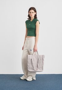 Szürke nylon bevásárlótáska két hosszú fogantyúval, cipzáras záródással és finom varrási részletekkel, zöld felsőt és bézs nadrágot viselő modell kezében.