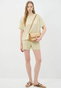 Licht beige kort mouwtop gecombineerd met licht beige shorts met een patroon. Het model houdt een tan crossbody tas met gevlochten details vast.