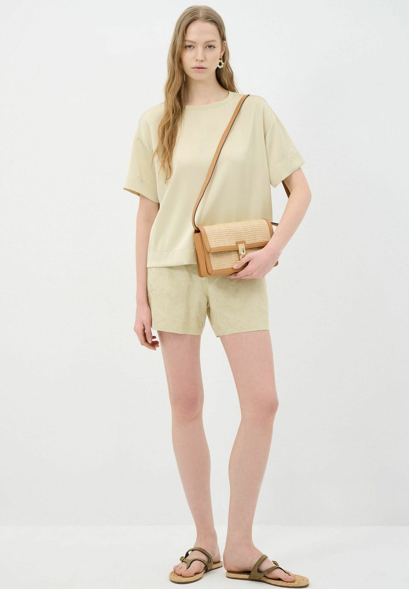 Licht beige kort mouwtop gecombineerd met licht beige shorts met een patroon. Het model houdt een tan crossbody tas met gevlochten details vast.
