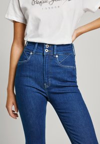 Jeans de mezclilla azul de cintura alta con dos bolsillos frontales, costuras sutiles y cierre de dos botones. Presenta un ajuste elegante y una textura suave.