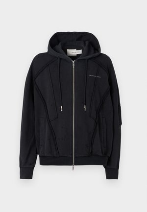 Črna zip-up kapuca iz mehkega blaga, z nastavljivo kapuco, stranskimi žepi in teksturiranimi paneli spredaj. Groba šivanka poudarja detajle.