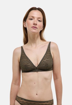 Femme portant un ensemble de lingerie transparente vert olive assorti avec des motifs de rayures ondulées sur un fond clair uni.