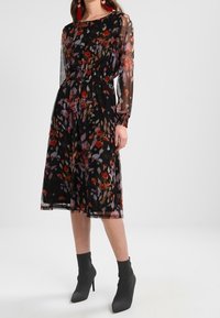 Černé květinové šaty s dlouhými průsvitnými rukávy, elastickým pasem a vrstvenou látkou. Mají vícebarevný květinový vzor a délku po kolena.