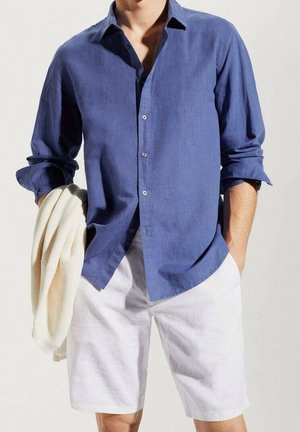 Camicia di lino blu con una vestibilità morbida, chiusura con bottoni davanti e maniche arrotolate, abbinata a pantaloni corti di lino bianchi. Maglione leggero appoggiato sul braccio.
