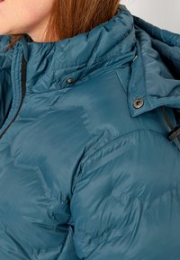 Puffet turkis jakke med høj krave, lynlås og trykknapper. Stoffet ser skinnende ud med en glat tekstur og blød isolering.