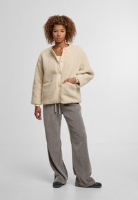 Kobieta z kręconymi włosami, ubrana w beżową polarową kurtkę, kremową bluzkę, luźne szare spodnie i ciemne buty, stoi na tle jasnego, jednolitego tła.