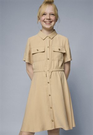 Vestido beige de manga corta hecho de una tela suave, con botones en la parte delantera, dos bolsillos en el pecho y una cintura con cordón para un ajuste ajustable.