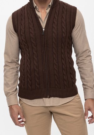 Mann trägt eine braune kabelgestrickte ärmellose Reißverschlussweste über einem beigen Hemd mit Knöpfen und hellbraunen Hosen, steht mit entspannten Händen an den Seiten.